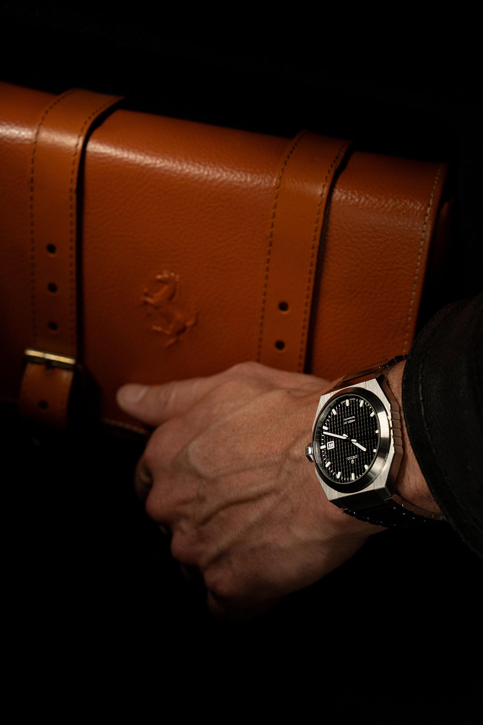 Hand holding a brown leather briefcase with a watch on a dark background. Model DWISS A1- using a Swiss made automatic movement La Joux-Perret G100 Soigné with 68 hours power reserve and white superLumiNova blocks