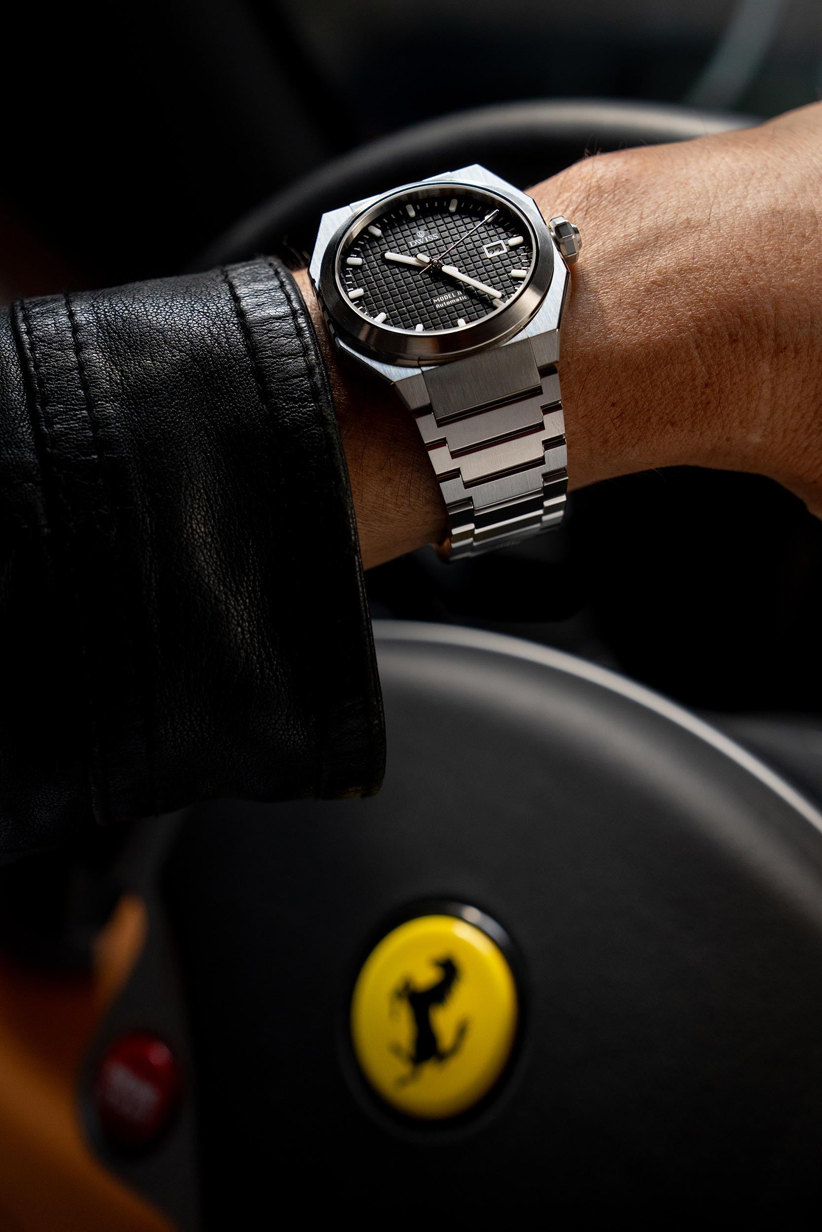 Person wearing a silver watch with a black face, sitting in a car with a Ferrari steering wheel. Model DWISS A1- using a Swiss made automatic movement La Joux-Perret G100 Soigné with 68 hours power reserve and white superLumiNova blocks