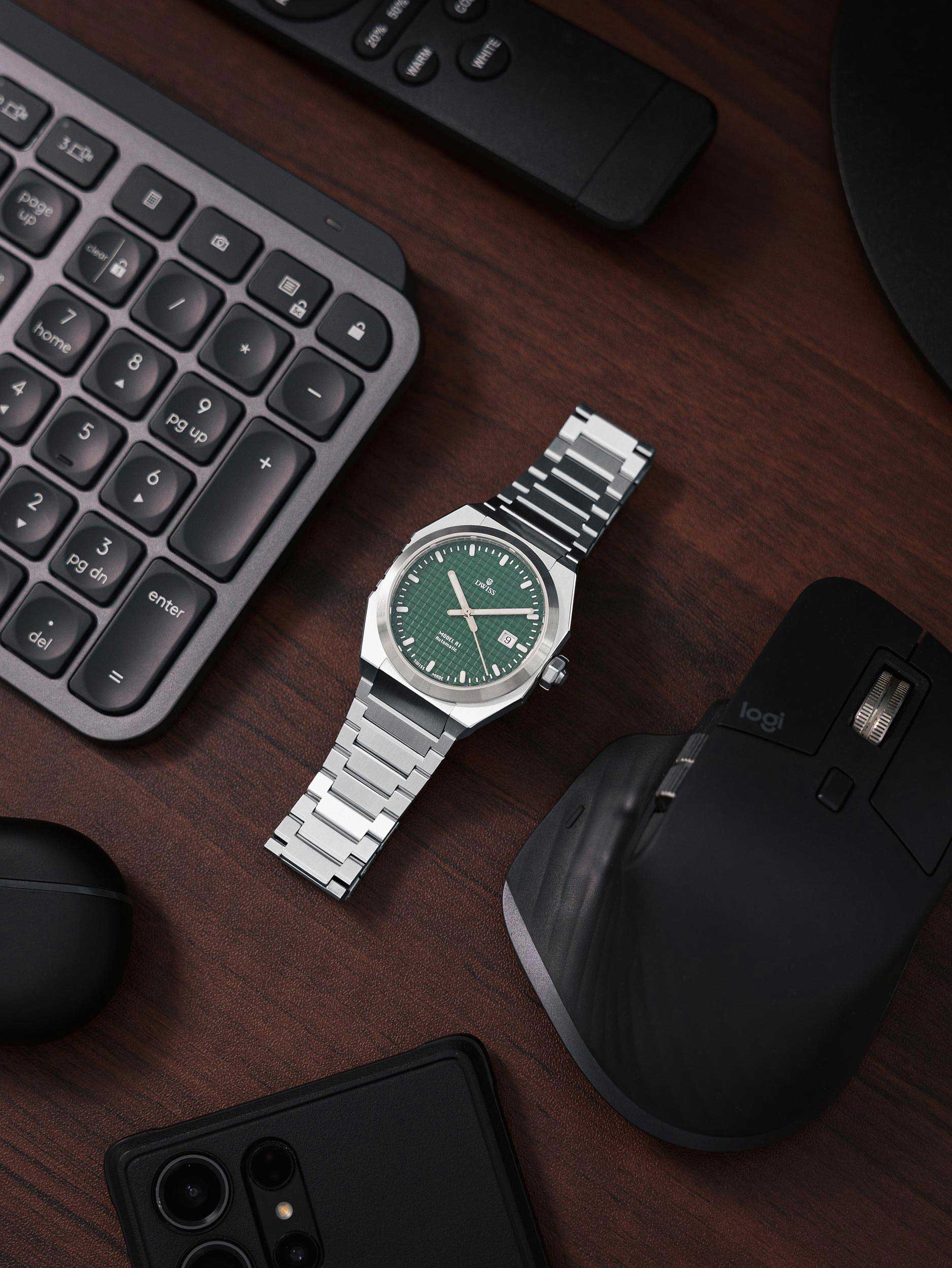 Silver watch with green face on a wooden desk with keyboard, mouse, and phone. Model DWISS A1- using a Swiss made automatic movement La Joux-Perret G100 Soigné with 68 hours power reserve and white superLumiNova blocks