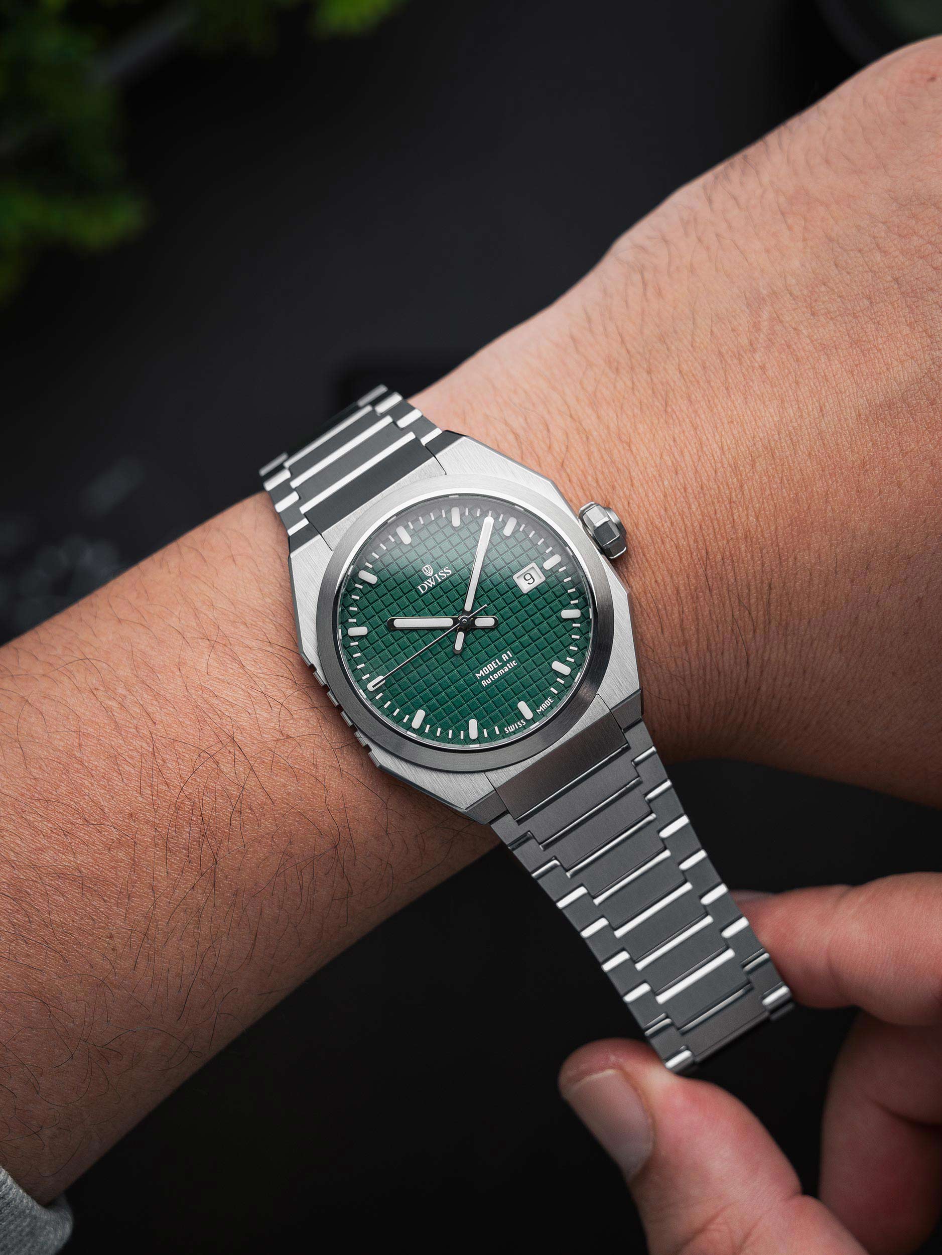 Silver watch with a green face on a wrist against a dark background. Model DWISS A1 - using a Swiss made automatic movement La Joux-Perret G100 Soigné with 68 hours power reserve and white superLumiNova blocks