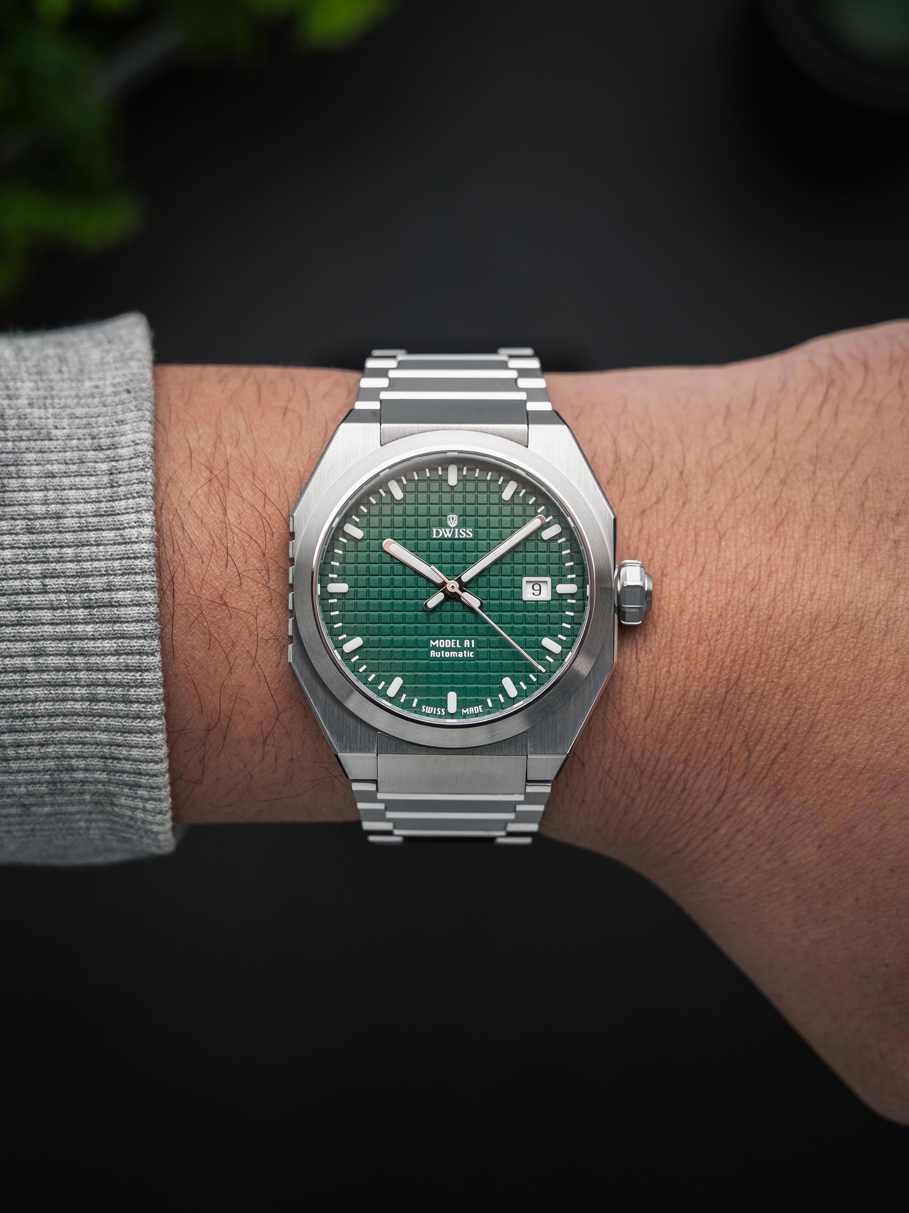 Silver watch with green dial on a wrist against a dark background. Model DWISS A1- using a Swiss made automatic movement La Joux-Perret G100 Soigné with 68 hours power reserve and white superLumiNova blocks