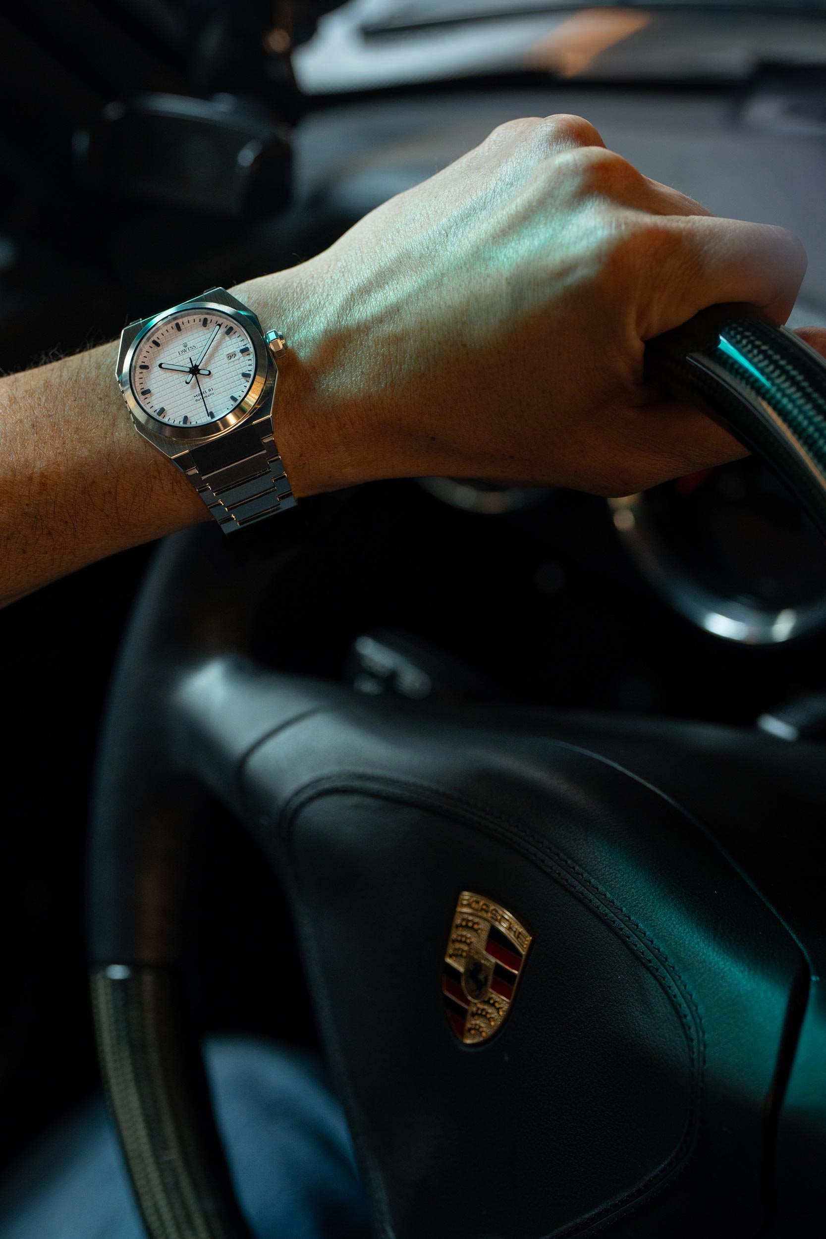 Hand on steering wheel of a car with a watch, Porsche emblem visible. Model DWISS A1- using a Swiss made automatic movement La Joux-Perret G100 Soigné with 68 hours power reserve and white superLumiNova blocks