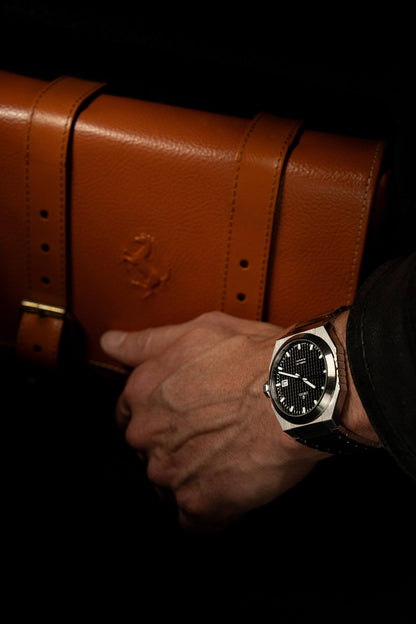 Hand holding a brown leather briefcase with a watch on a dark background. Model DWISS A1- using a Swiss made automatic movement La Joux-Perret G100 Soigné with 68 hours power reserve and white superLumiNova blocks