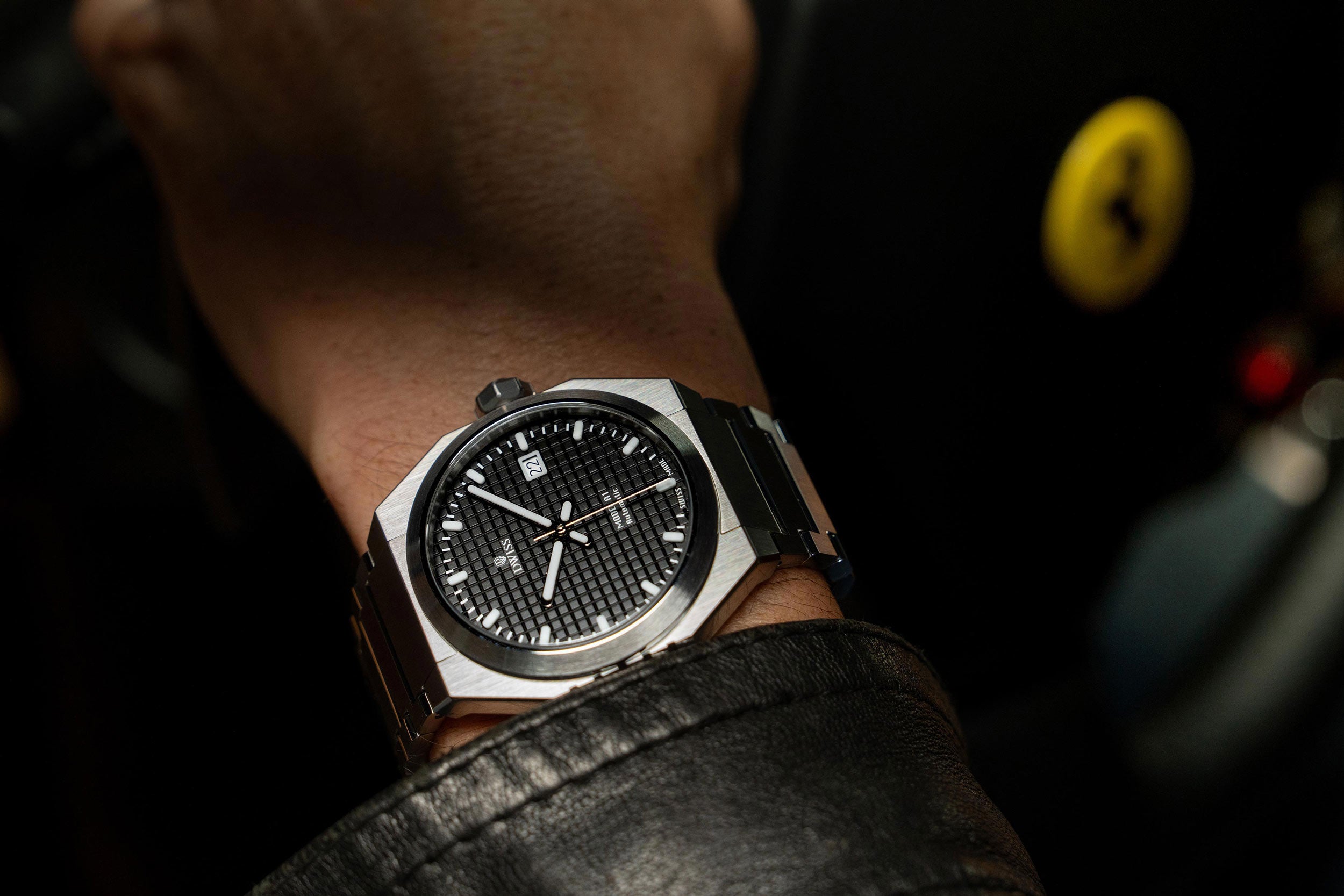 Watch on a wrist with a blurred background featuring a FErrari logo. Model DWISS A1- using a Swiss made automatic movement La Joux-Perret G100 Soigné with 68 hours power reserve and white superLumiNova blocks