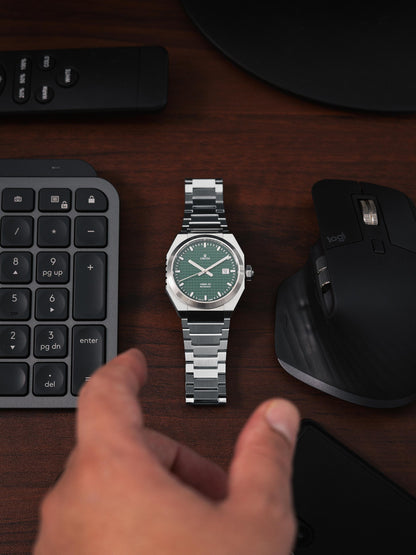 Silver watch with green face on a desk next to a keyboard and mouse. Model DWISS A1- using a Swiss made automatic movement La Joux-Perret G100 Soigné with 68 hours power reserve and white superLumiNova blocks