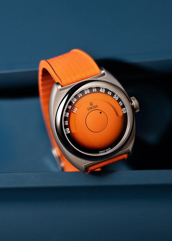 DWISS S1 Wandering Hours Naranja con esfera de fume gradiente negro. Reloj de pulsera con una correa de naranja brillante y un dial de color naranja, situado en un fondo azul oscuro.El reloj tiene una caja de m eta l con un bisel circular y un logotipo de la marca DWISS visible en el dial.