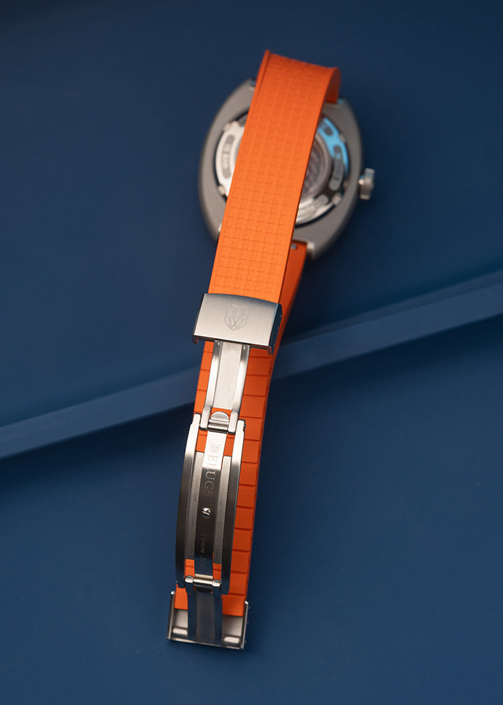 DWISS S1 Wandering Hours Naranja con esfera de fume gradiente negro. Cierre de una correa de reloj con una banda de goma de color naranja brillante y pulido de plata-tone m eta l hardware, unido a una caja redonda de plata reloj contra un fondo azul oscuro.