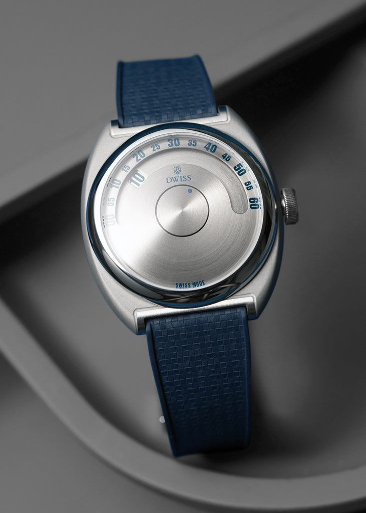 DWISS S1 Wandering Horas esfera circular cepillada, con una caja ovalada de plata cepillada, una correa de textura azul, y un dial de plata minimalista con marcas azules, fotografiado en una superficie gris oscura.