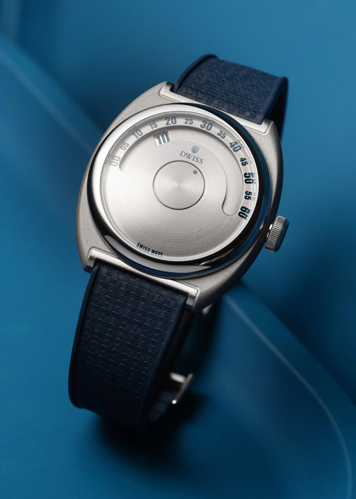 DWISS S1 Wandering Horas esfera circular cepillada, un plano de un reloj suizo con una caja de plata cepillada, un dial blanco y una correa azul de textura oscura, descansando en un fondo azul.