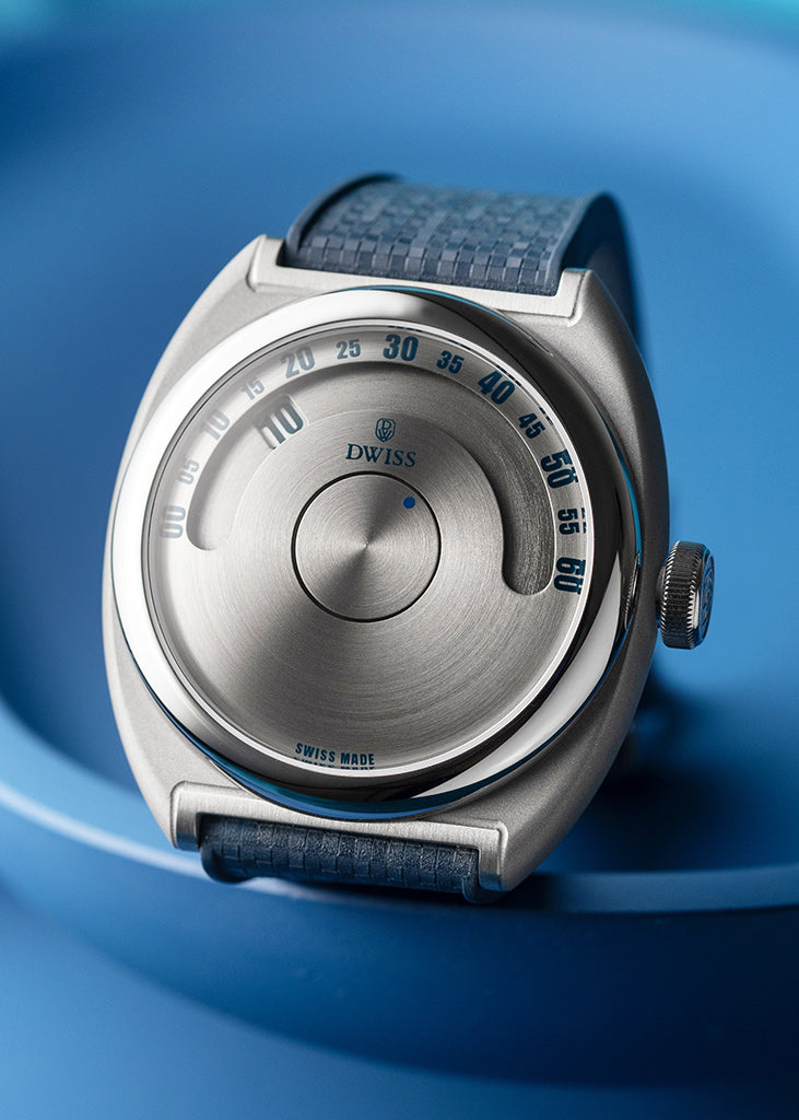 DWISS S1 Wandering Horas esfera circular cepillada, Un plano de un reloj suizo con una caja de acero inoxidable cepillado y esfera, con una correa azul texturada, enmarcada en un fondo azul coincidente.